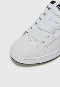 Zapatilla de piel blanca con puntera perforada, cordones blancos planos y una suela de goma texturizada. Presenta un pequeño logo de la marca en el costado.