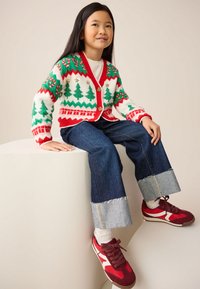 Pleteni jopič z vzorci prazničnih dreves in okraskov v rdeči, zeleni in beli barvi; kombiniran s temno modrimi kavbojkami z raztrganimi robovi in rdečimi supergami.