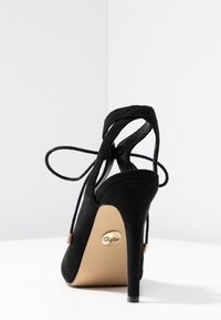 Czarne zamszowe szpilki z paskami na kostkę związanymi w kokardę, z widoczną beżową podeszwą z logo "Buffalo" i oznaczeniem rozmiaru 37.