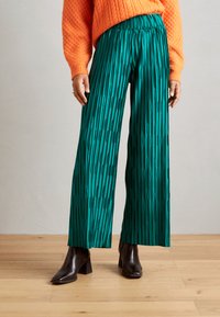 Pantalones anchos de cintura alta en verde con pliegues y rayas verticales, hechos de un tejido ligero, combinados con botas de tacón negro y un suéter texturizado naranja.