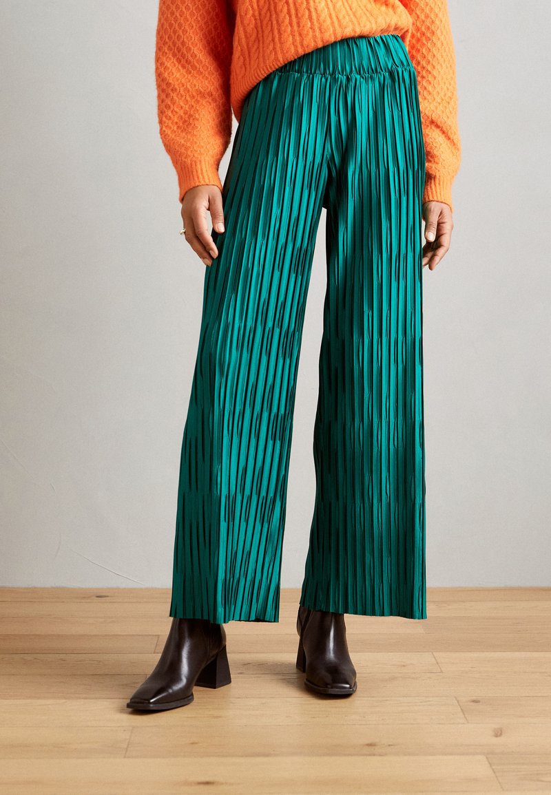 Pantalones anchos de cintura alta en verde con pliegues y rayas verticales, hechos de un tejido ligero, combinados con botas de tacón negro y un suéter texturizado naranja.