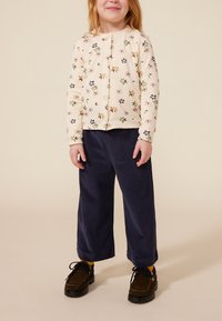 Crèmekleurig vest met bloemenprint, marineblauwe corduroy broek met wijde pijpen, en donkerbruine schoenen. Het vest heeft knopen en lange mouwen.