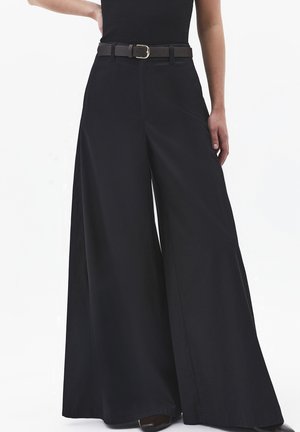 Persona que lleva pantalones negros de corte ancho y cintura alta con un cinturón negro y una camiseta negra, de pie con una mano en la cadera y la otra relajada.