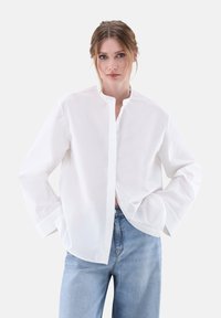 Camicia bianca in cotone con colletto alla cinese, maniche lunghe, chiusura frontale con bottoni e vestibilità ampia, abbinata a jeans in denim blu chiaro.