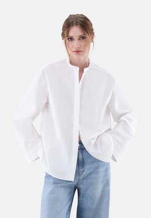 Camicia bianca in cotone con colletto alla cinese, maniche lunghe, chiusura frontale con bottoni e vestibilità ampia, abbinata a jeans in denim blu chiaro.