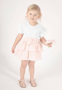 Biały top o fakturze z falbankami na rękawach, zestawiony z różową, wielowarstwową spódnicą z kokardą. Dziecko trzyma kwiat i nosi sandały w wzory.