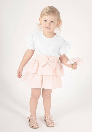 Biały top o fakturze z falbankami na rękawach, zestawiony z różową, wielowarstwową spódnicą z kokardą. Dziecko trzyma kwiat i nosi sandały w wzory.