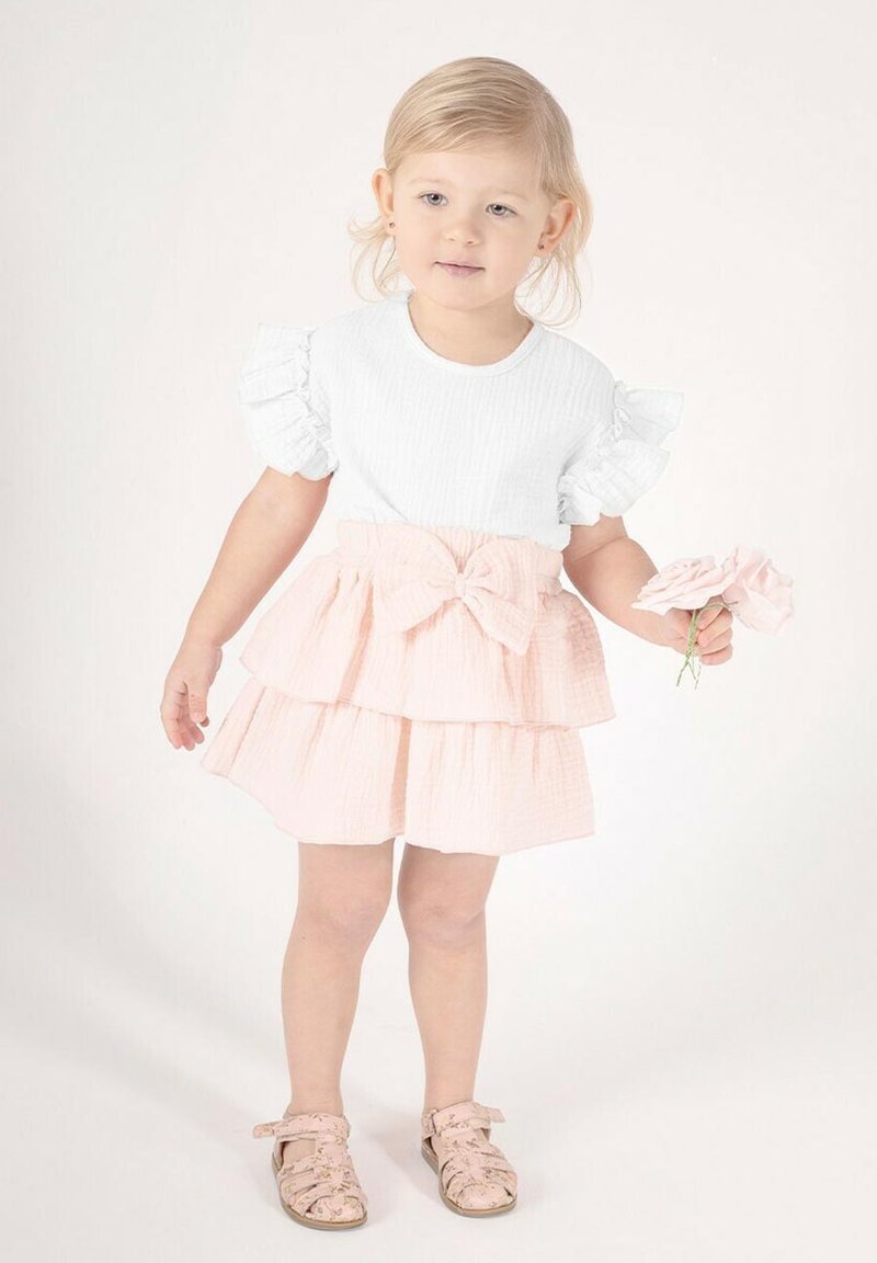 Biały top o fakturze z falbankami na rękawach, zestawiony z różową, wielowarstwową spódnicą z kokardą. Dziecko trzyma kwiat i nosi sandały w wzory.