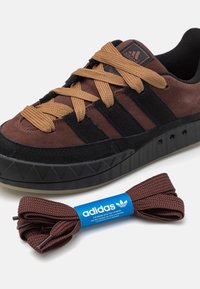 Hnědé semišové tenisky s černými akcenty a trojúhelníkovým texturovým detailem na podešvi. Zahrnují hnědé tkaničky s modrou nášivkou adidas.