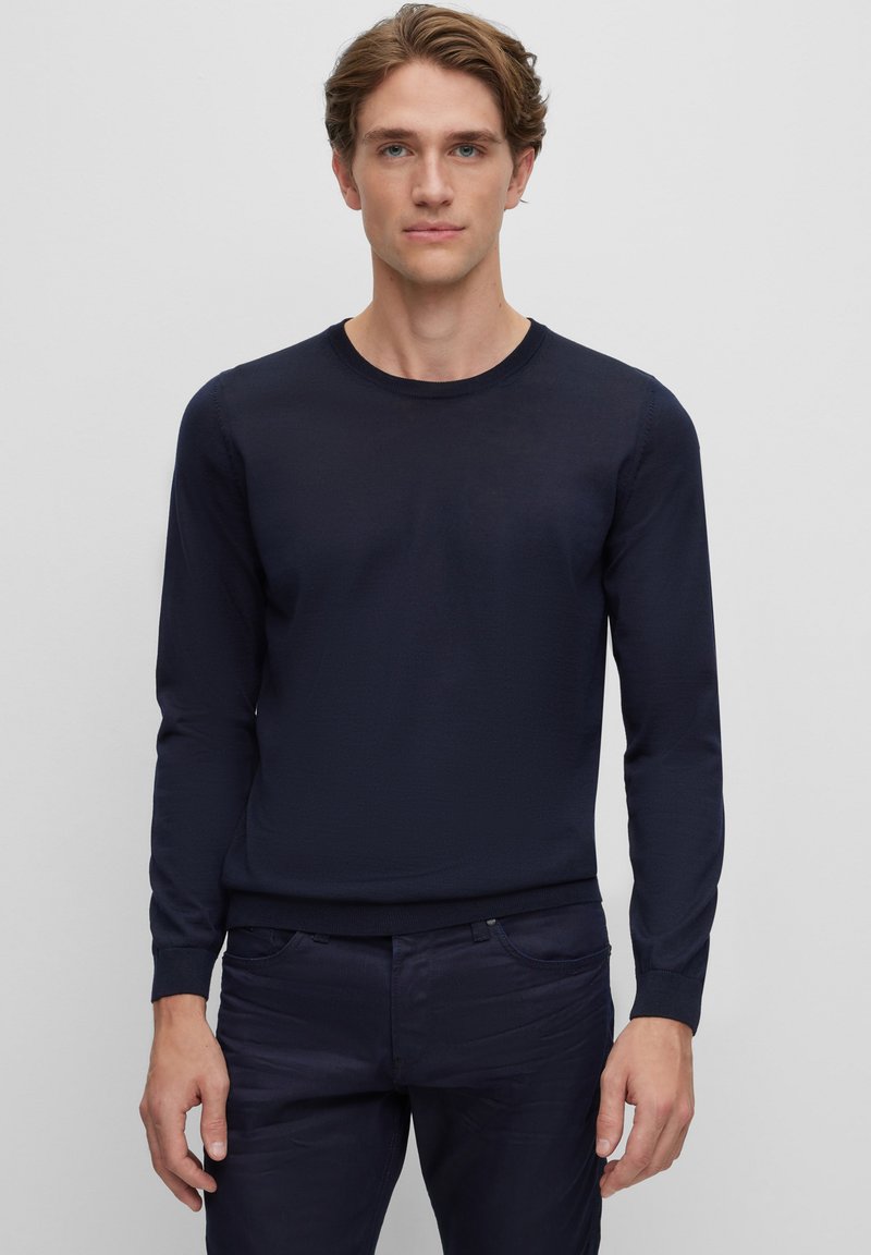 Pulover albastru închis, cu mâneci lungi și guler rotund, cu manșete din tricot. Textură moale, design simplu, asortat cu pantaloni negri.