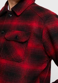 Chemise en flanelle à carreaux rouges et noirs avec col pointu, deux poches avant et boutons noirs. Texture douce avec une apparence chaleureuse.