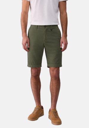 Uomo con pantaloncini verde oliva fino al ginocchio, t-shirt bianca e sneaker in camoscio beige, in piedi davanti a uno sfondo semplice.