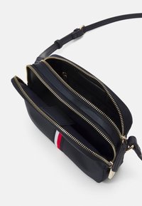 Schwarze Leder-Crossbody-Tasche mit goldenen Reißverschlüssen, zwei Fächern und einem roten und weißen Streifen-Detail auf der Vorderseite. Verstellbarer Riemen enthalten.