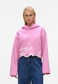 Rosa, verkürzte Kapuzenjacke aus weichem Stoff, mit einem weißen, geschwungenen "Karl" am Saum und einer vorderen Kängurutasche. Lange, weite Ärmel.
