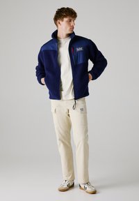 Marine fleece jack met een blauwe nylon schouderstuk, staande kraag, rits borstzak, gecombineerd met crèmewitte cargobroek en sneakers.