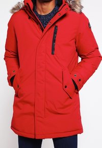 Parka isolée rouge avec capuche amovible doublée en fourrure, poche zippée sur la poitrine, deux poches à l'avant et une texture solide.