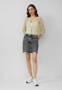 Lichtgroene, gebreide mesh vest over een beige tanktop; gecombineerd met een zwarte denim rok met een rafelige zoom en witte sneakers.