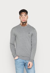 Grijze lange mouwen sweater met een ronde hals en geribbelde manchetten. Klein geborduurd logo op de linkerkant van de borst. Gemaakt met lichtblauwe jeans.