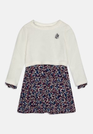 Langærmet børnekjole med hvid top med et lille våbenskjold og et flerfarvet nederdel med blomsterprint i marineblå, rød og hvid.