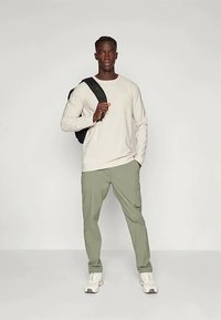 Bărbat în picioare cu un rucsac negru pe umăr, îmbrăcat cu o cămașă bej cu mânecă lungă, pantaloni cargo verzi și adidași albi pe un fundal simplu.