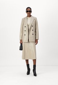 Beige Blazer aus Kunstleder mit doppelreihigen Knöpfen, kombiniert mit einem passenden Rock. Schwarze Accessoires und eckige Sonnenbrillen runden den Look ab.