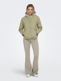 Kvinde stående iført quiltet olivengrøn jakke over grå hættetrøje, beige flarebukser og hvide sneakers, hænderne i jakkens lommer.