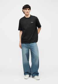 Νέος με κατσαρά μαλλιά, ντυμένος με μαύρο T-shirt, φαρδιά μπλε τζιν και λευκά αθλητικά παπούτσια, στέκεται με ένα χέρι πίσω από την πλάτη του.