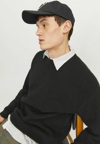 Schwarzer, gestrickter Pullover aus strukturiertem Stoff, über einem weiß-schwarz gestreiften Hemd mit Kragen getragen. Schwarze Baseballkappe. Sitzt auf einem Stuhl.
