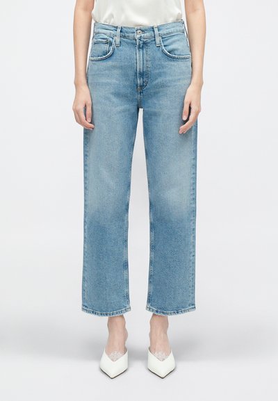 Jean en denim bleu clair, taille haute, avec des jambes droites et un léger délavage, dotés de poches avant et de passants de ceinture, associés à des chaussures blanches.