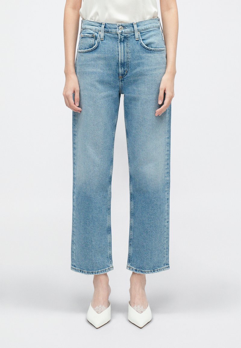 Jeans din denim de culoare albastră deschisă, cu talie înaltă, picioare drepte și ușoară decolorare, dotate cu buzunare frontale și bucle pentru curea, stilizate cu �încălțăminte albă.