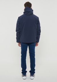 Veste imperméable navy avec un col montant et une capuche réglable, associée à un jean bleu et des baskets noires. Design simple, matériau fonctionnel.