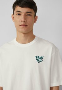 Jeune homme aux cheveux courts et foncés portant un t-shirt blanc avec le texte "Field Trip" en vert sur la gauche de la poitrine, fond gris neutre.