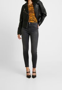 Chaqueta de cuero negra estilo motero con detalles acolchados, top a rayas naranja y negro, jeans gris ajustados y tacones negros de punta afilada.