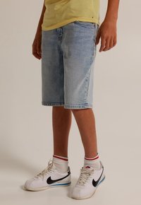 Světle modré džíny šortky s uvolněným střihem, obsahující přední kapsy a třepený lem. Noseny s bílými teniskami a pruhovanými ponožkami.