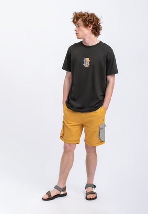 T-shirt nero a maniche corte con una grafica di una casa colorata, abbinato a pantaloni cargo gialli con accenti grigi e molteplici tasche. Includendo sandali.