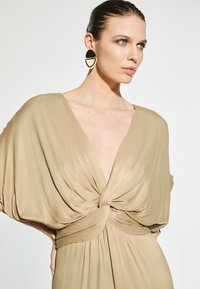 Beige wrap dress with a deep V-neck and twisted bodice detail. Features wide sleeves and a flowing silhouette. Black and white circular earrings.