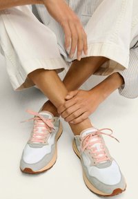 Las zapatillas presentan ante gris claro, malla blanca y suaves acentos en rosa. Tienen una forma redondeada y una lengüeta acolchada; los cordones son de un rosa pálido, con una suela beige.