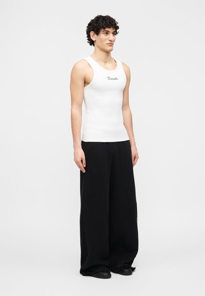 Jeune homme debout portant un haut blanc ajusté sans manches avec le texte "Endorphin" et un pantalon noir à jambes larges sur un fond blanc uni.