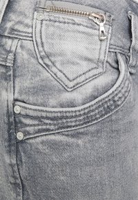 Jeans de denim gris claro que presentan un bolsillo con cremallera, costuras distintivas y cierres de botón. La tela tiene una textura desgastada con un corte clásico.