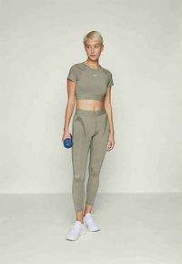 Kvinne i grågrønn treningstopp og leggins holder en blå hantel, stående mot en enkel lys bakgrunn iført hvite joggesko.