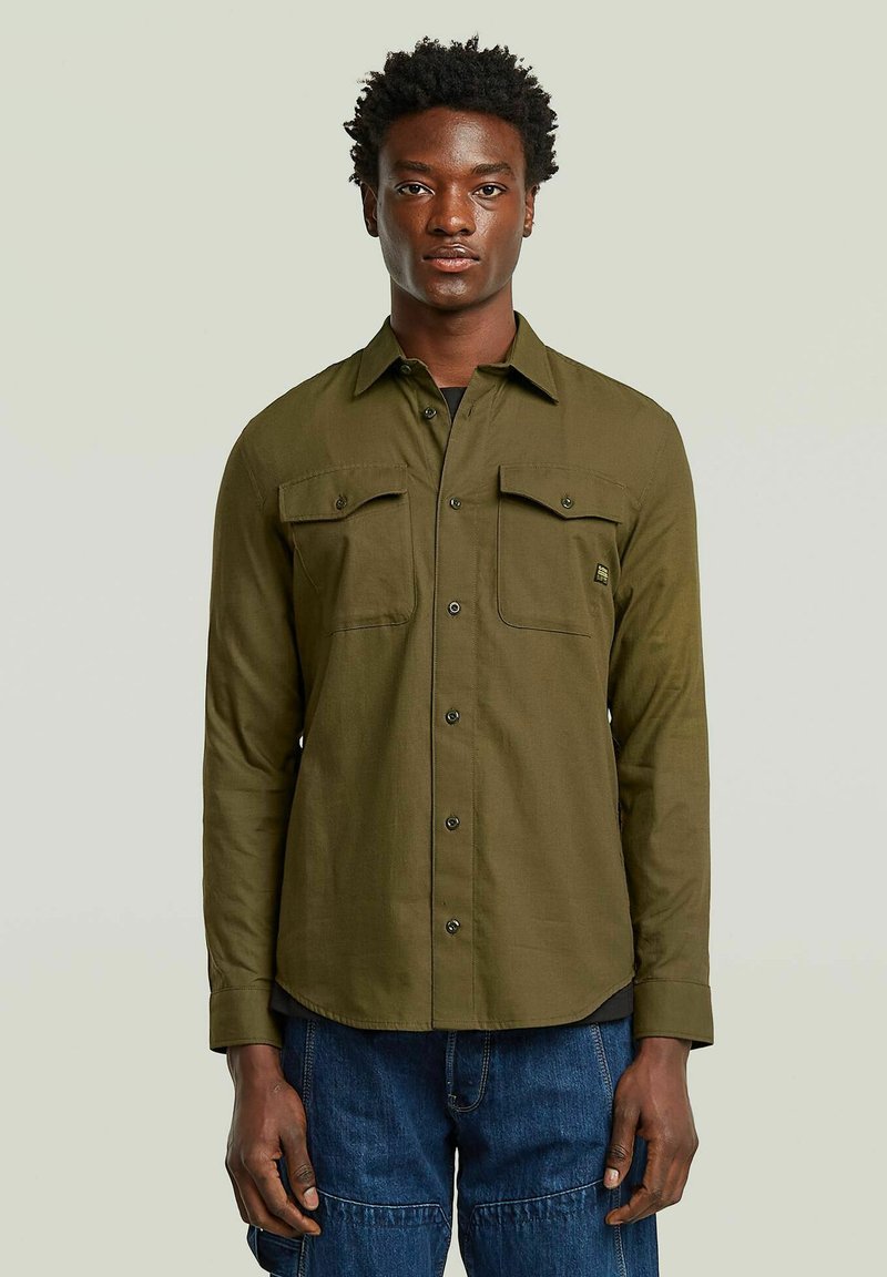 Chemise à manches longues vert olive avec deux poches poitrine et boutons noirs. Présente un col pointu et une coupe droite à l'ourlet.
