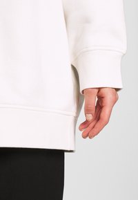 Sudadera blanca con puño y dobladillo acanalados. El diseño es sencillo con una textura suave, combinada con un pantalón negro visible en la parte inferior.