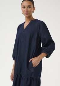 Námornícky denimový šaty s výstrihom do V, nadýchanými rukávmi a elastickými manžetami. Obsahujú stupňovitý lem a bočné vrecko pre funkčnosť.