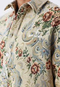 Camicia a maniche corte con motivo floreale beige, caratterizzata da accenti rossi e blu, tessuto testurizzato e un piccolo logo sul petto.