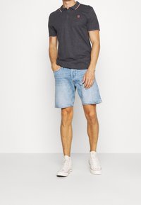 Homme portant un polo gris foncé avec des bordures roses, un short en denim léger bleu, et des baskets montantes blanches, se tenant contre un fond blanc.