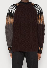 Maglione a maglia spessa con corpo marrone scuro, motivi a cavo diagonale e maniche a zig-zag multicolori nei toni del grigio, bianco e arancione.