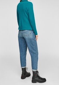 Haut à manches longues bleu sarcelle avec col, associé à un jean bleu clair à coupe décontractée. Bottes noires à lacets avec détails texturés et semelles épaisses.