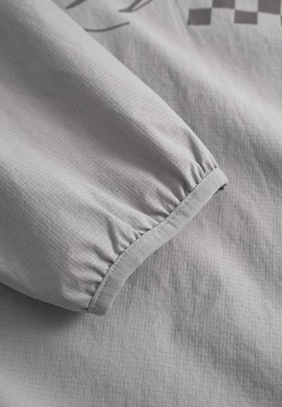 Tissu gris clair avec un motif texturé subtil. La manche froncée présente un poignet cousu, mettant en valeur des éléments de design simples.