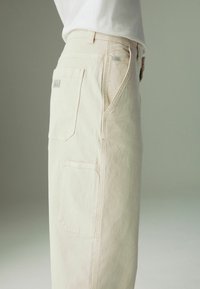Beige cargo pants featuring side pockets and a logo patch, made of textured fabric, with a straight fit.