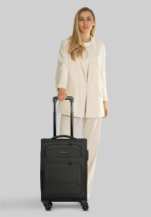 Vrouw in licht pak die het handvat vasthoudt van een zwarte koffer op wielen met het label "SMARTBOX" tegen een effen grijze achtergrond.